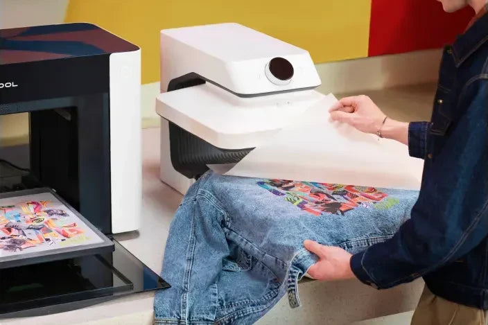 Peeling a DTF transfer film off a denim jacket after pressing, with the xTool O1 DT+UV beside it