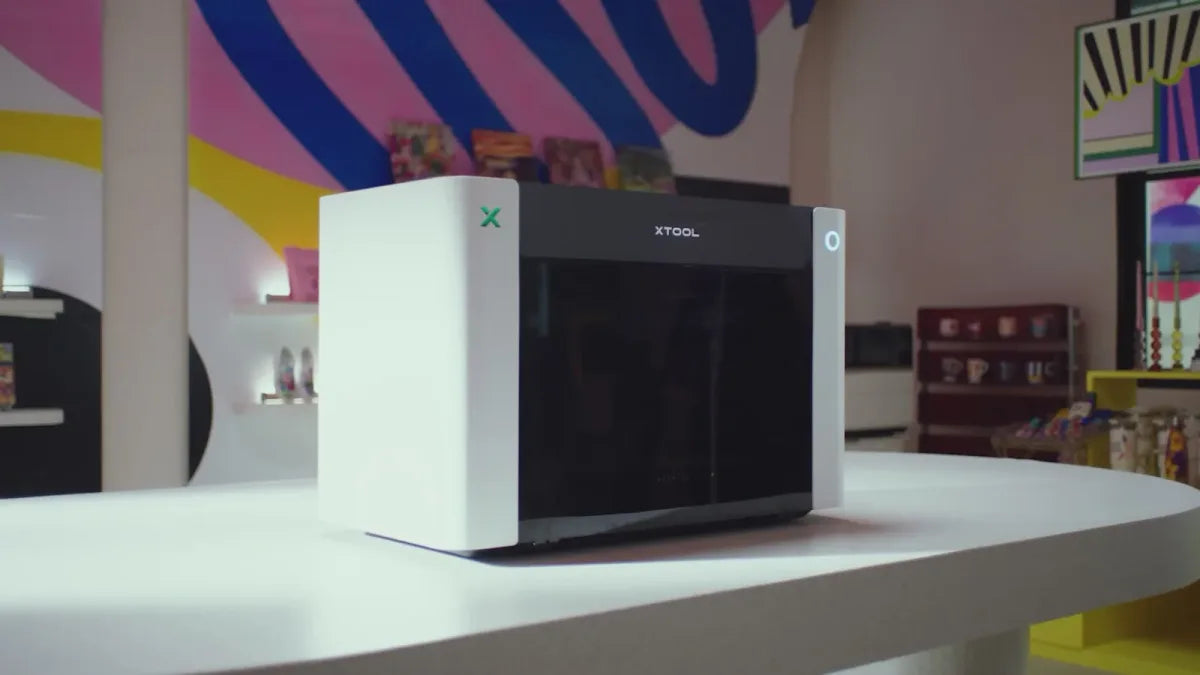 The xTool O1 Omni Printer on a shop counter, with printed mugs and art on the shelves behind it
